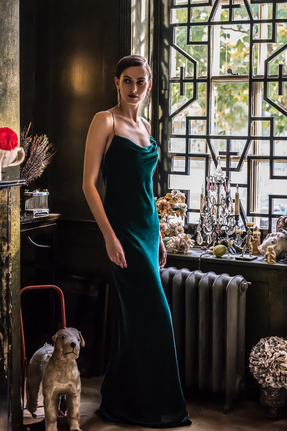 A woman in a green silk velvet maxi dress with a deep neckline and spaghetti straps, standing in a room with decorative items and a dog on a chair.