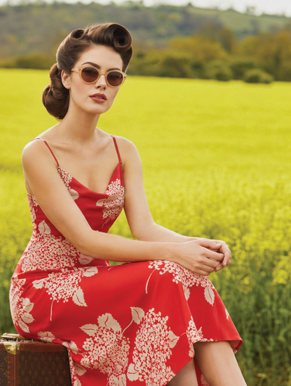 A woman in a red floral midi dress and round sunglasses sits on a vintage suitcase in a bright yellow field.